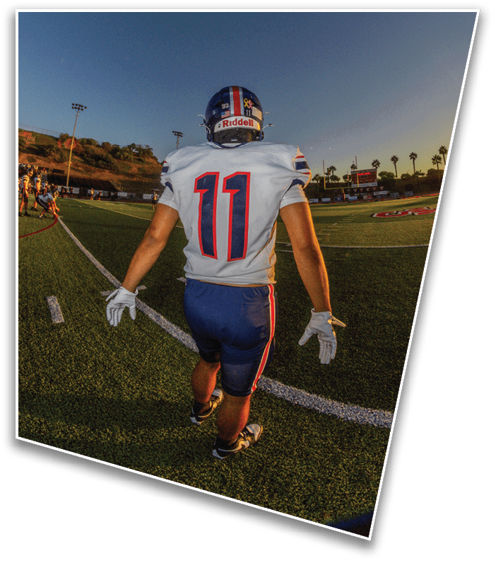 A person wearing a football uniform with the number 11 is walking on a football field during sunset with other players and stadium lights visible in the background. Description generated by AI