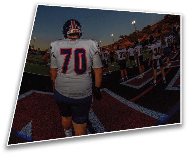 A person wearing a football helmet and jersey stands on a field under a darkening sky with stadium lights illuminating the scene. Description generated by AI