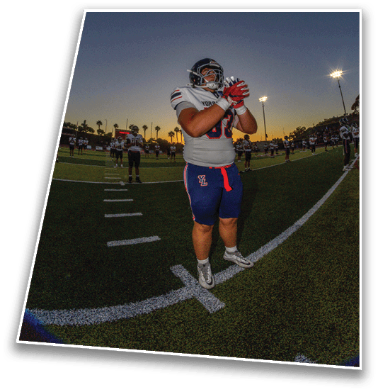 A person in football gear stands on a field at dusk with other players and stadium lights in the background. Description generated by AI