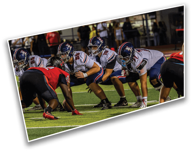 Several people in football uniforms are positioned on a field facing each other, preparing for a play during a game. Description generated by AI