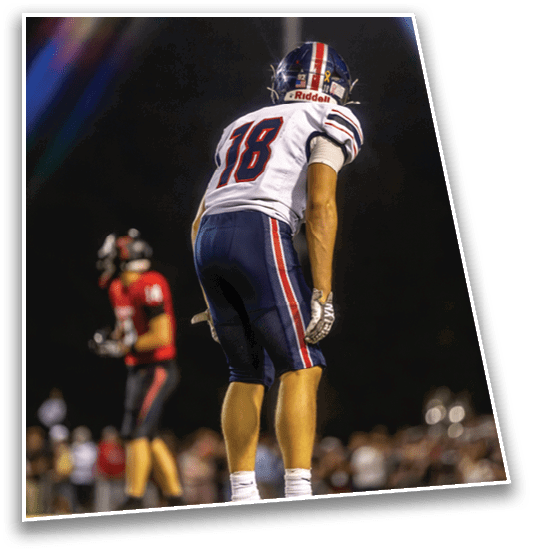 A person wearing a white football jersey with the number 18 and blue pants is standing on a field facing away, while another person in a red and black uniform is blurred in the background. Description generated by AI