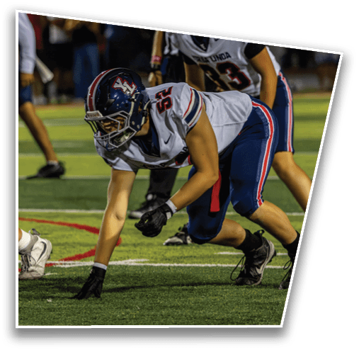 People wearing athletic gear and cleats are crouched in a ready position on a grassy sports field, likely preparing for a play in a football game. Description generated by AI