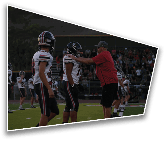 A person in a red shirt and cap appears to be coaching or instructing several people in football uniforms on a field with spectators in the background. Description generated by AI