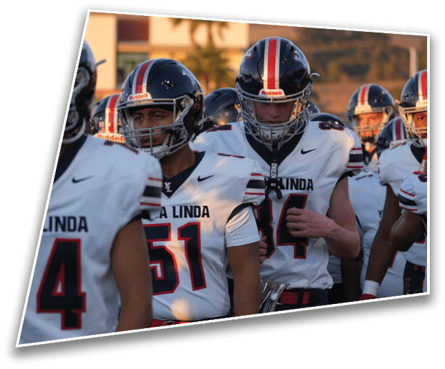 People wearing football helmets and uniforms stand together outdoors with a hill and palm trees in the background. Description generated by AI