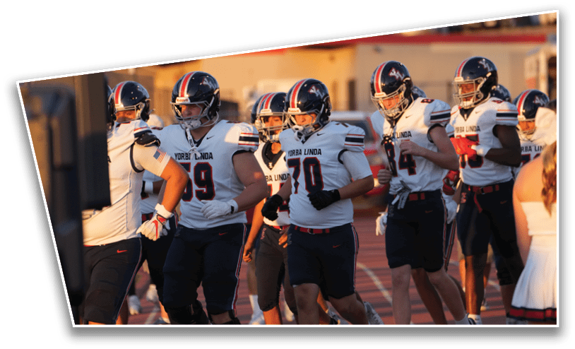 A group of football players in white and navy uniforms with helmets are standing together on a field near a cheerleader in a white outfit during a game or practice. Description generated by AI