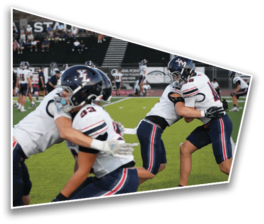 Two pairs of people in football uniforms and helmets are engaged in blocking drills on a green field with other players and spectators in the background. Description generated by AI