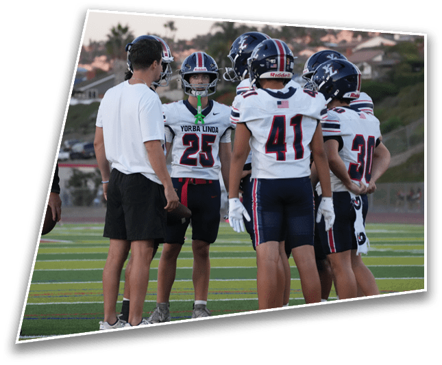 People wearing football uniforms and helmets are standing on a field while a person in a white shirt and black shorts appears to be talking to them. Description generated by AI