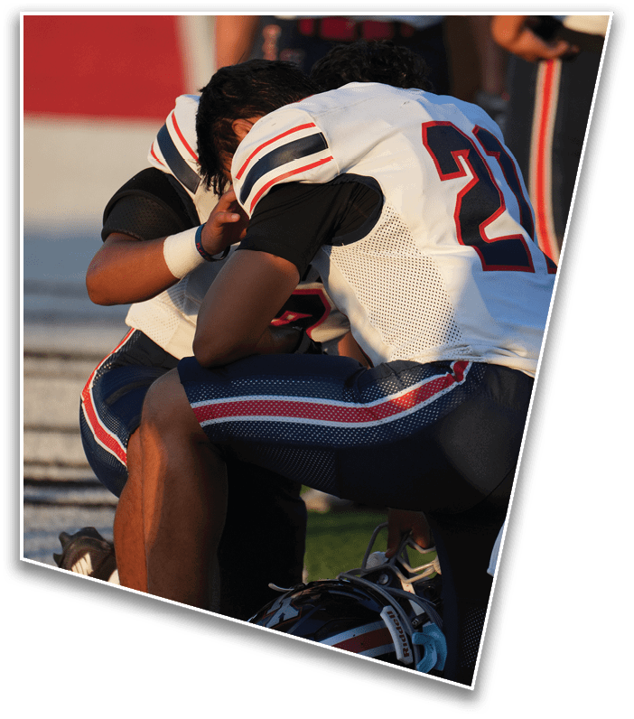 Two people in football uniforms kneel on the field with one resting their head on the other's shoulder and a helmet on the ground nearby. Description generated by AI