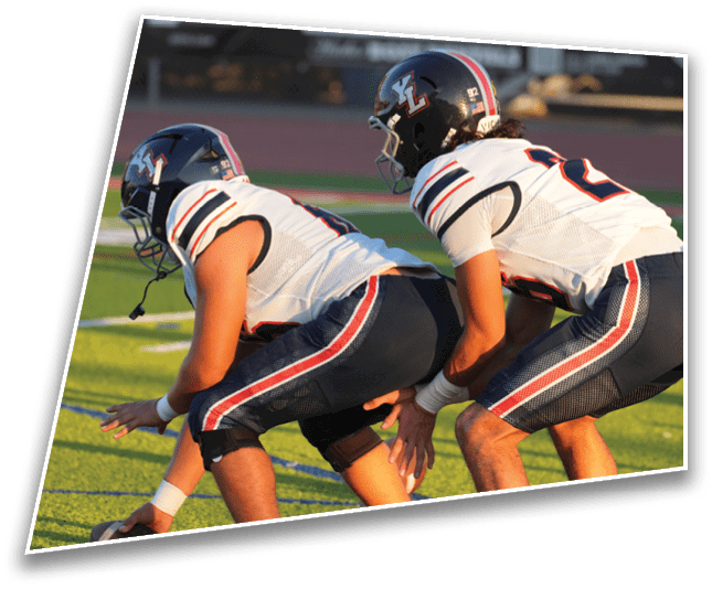 Two people wearing white football uniforms and helmets are positioned on a football field, appearing ready for a play. Description generated by AI