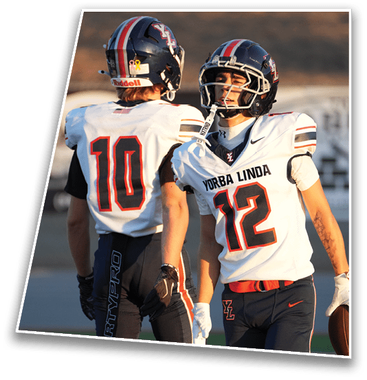 Two people wearing football uniforms with helmets are on a field, one holding a football and the other facing away. Description generated by AI
