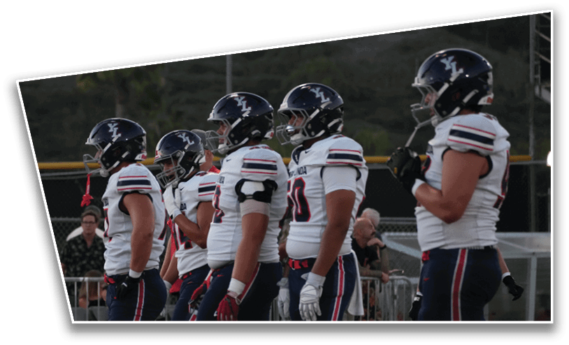Five people in football uniforms and helmets stand in a line on a field with a track and fencing in the background. Description generated by AI