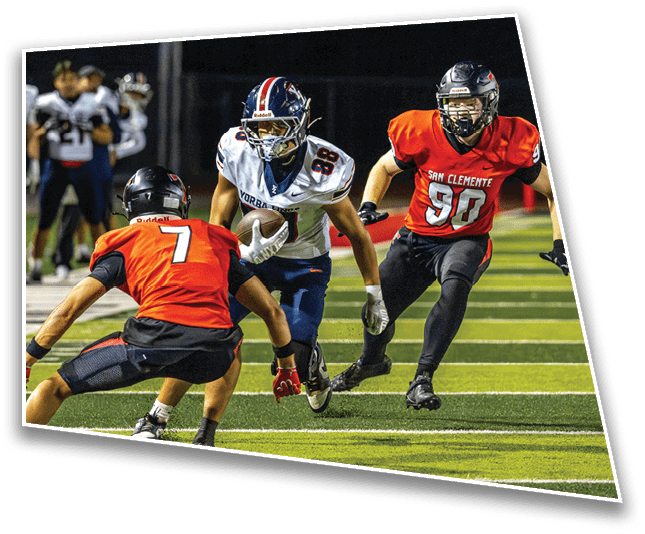 Three people in football uniforms are actively playing on a field with one person holding the ball while two others in red jerseys attempt to block or tackle. Description generated by AI