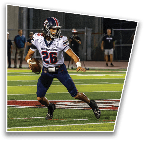 A person wearing a football uniform with the number 26 is running on a field while holding a football, with several people standing in the background. Description generated by AI
