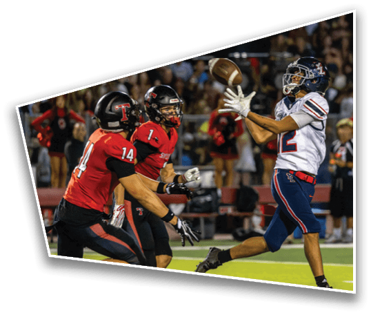 A person in a white and blue football uniform is reaching to catch a football while two people in red and black uniforms are running toward them on a football field. Description generated by AI