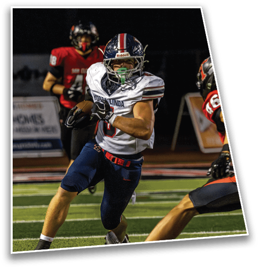 A person in a white and blue football uniform is running with the ball while two people in red and black football uniforms are approaching to tackle. Description generated by AI