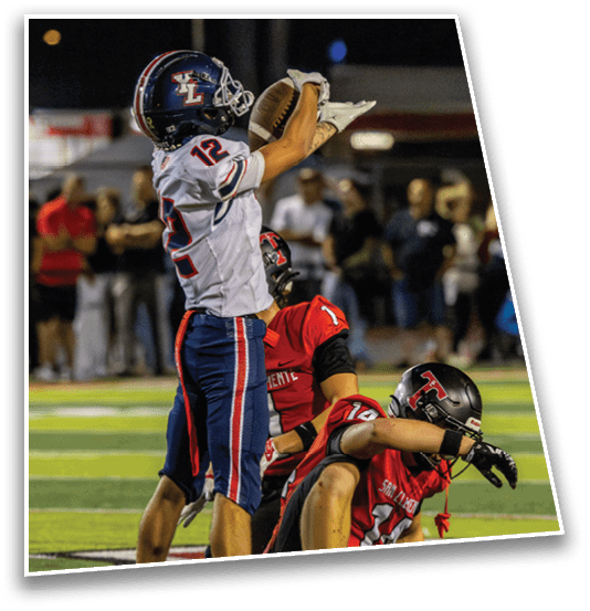 Two football players in red uniforms are on the ground while a player in a white and blue uniform stands nearby on a football field with spectators in the background. Description generated by AI