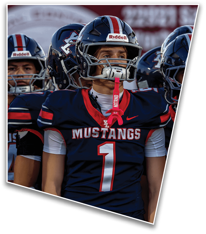 A person wearing a navy blue and red Mustangs football jersey with the number one and a helmet with a pink mouthguard hanging from the face mask stands next to another person in similar gear. Description generated by AI