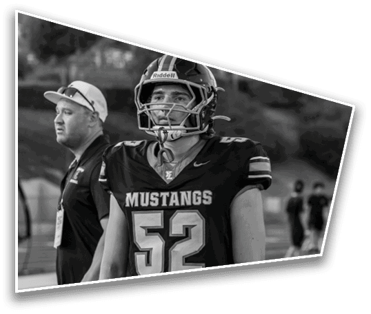 A person wearing a football helmet and Mustangs jersey stands next to another person wearing a cap and sunglasses on their head in an outdoor setting. Description generated by AI