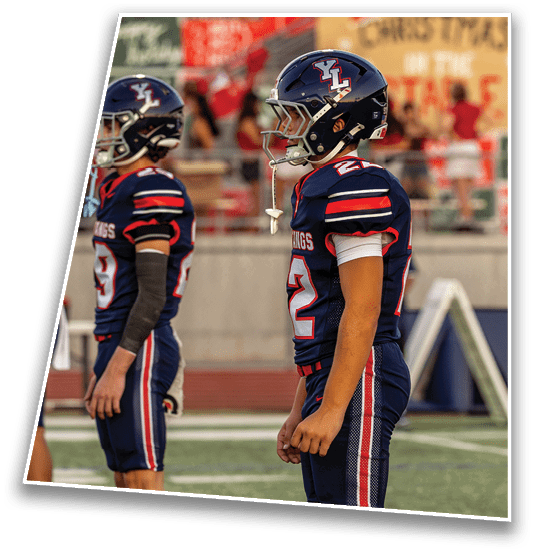 Two people in football uniforms and helmets stand on a field with a blurred crowd and a Christmas-themed sign in the background. Description generated by AI