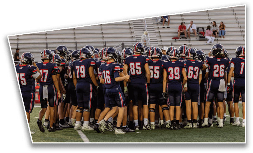 A group of football players in dark uniforms with helmets are huddled together on a field with empty bleachers in the background. Description generated by AI