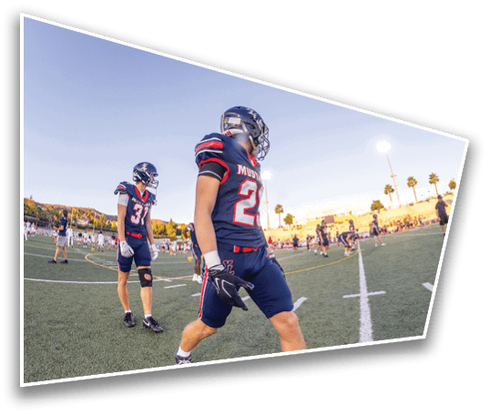 Two people in football uniforms and helmets walk on a field with other players and spectators in the background during a clear evening. Description generated by AI