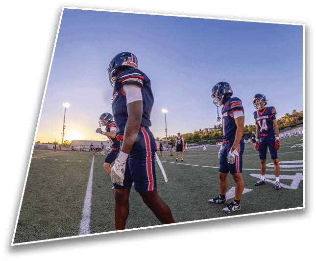 People wearing football uniforms are standing and walking on a field with artificial turf during sunset. Description generated by AI
