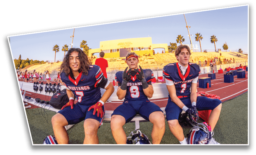 Three people in football uniforms sit on a bench on a sports field with a stadium and palm trees in the background during early evening. Description generated by AI