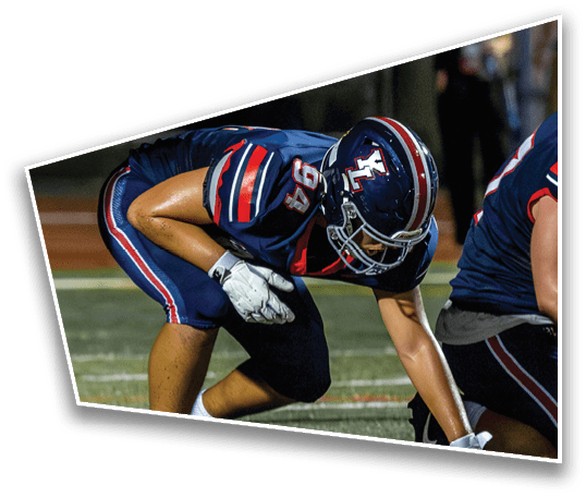 Two people in football uniforms and helmets are crouched on a field, preparing for a play. Description generated by AI