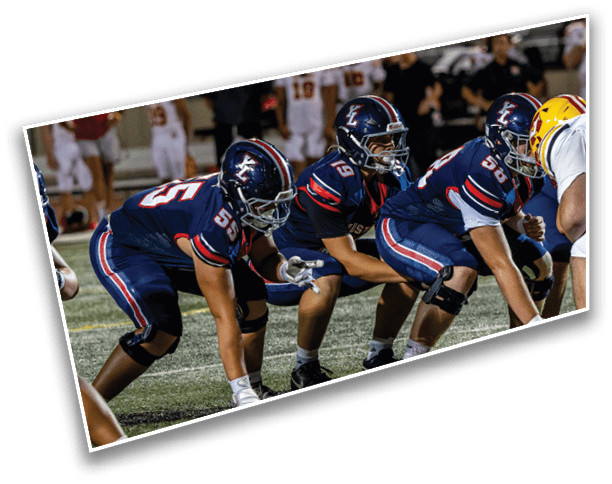 Three people in blue football uniforms are lined up on a field preparing for a play against an opposing player in a yellow helmet. Description generated by AI
