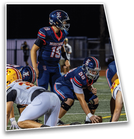 Two football players in navy blue uniforms with “Mustangs" on the jersey prepare for a play on the field while opposing players in white and yellow uniforms line up against them. Description generated by AI