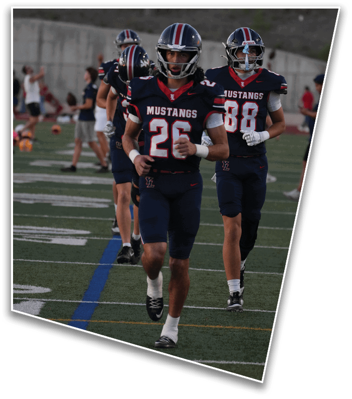 Three people in football uniforms with helmets are walking on a field with yard lines and a blue line visible. Description generated by AI