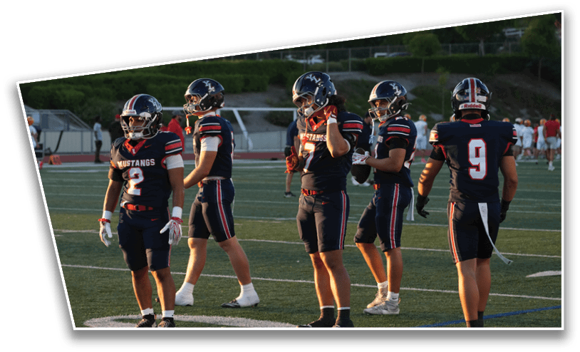 Five people wearing dark football uniforms with helmets stand on a sports field during a game or practice. Description generated by AI