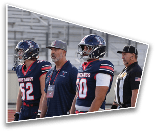 Two people in football uniforms and helmets stand next to two adults, one wearing a cap and a lanyard and the other in a referee uniform, on a sports field with empty bleachers in the background. Description generated by AI