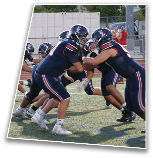 People wearing athletic uniforms and cleats are crouched in a ready position on a grassy sports field, likely preparing for a play in a football game. Description generated by AI