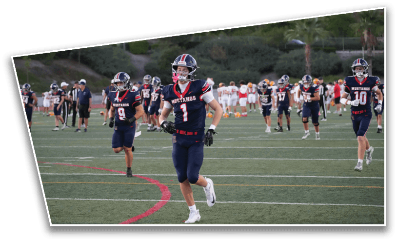 Several people wearing navy blue football uniforms with red and white accents are running on a green sports field with other players and spectators in the background. Description generated by AI