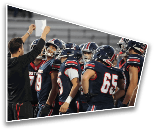 A group of football players in navy blue uniforms with red and white stripes are gathered around a coach who is holding up a piece of paper on a sports field. Description generated by AI