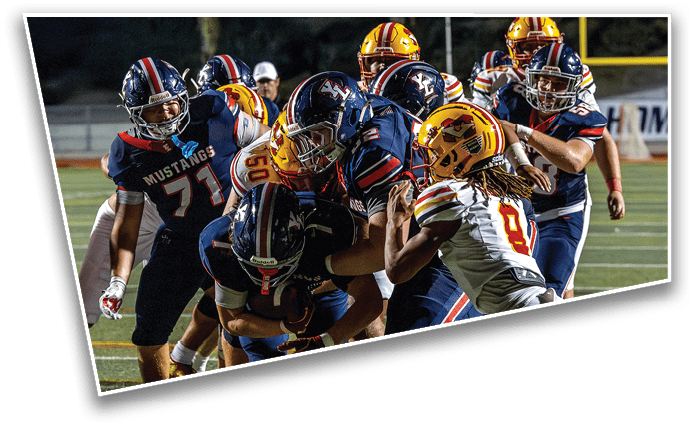 Several football players in helmets and uniforms are engaged in a tackle on a well-lit field during a game. Description generated by AI