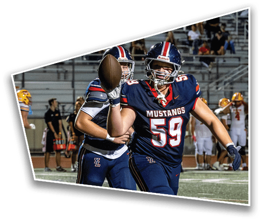 Two people wearing football helmets and uniforms are on a field with one person holding a football in mid-throw during a game with spectators in the background. Description generated by AI