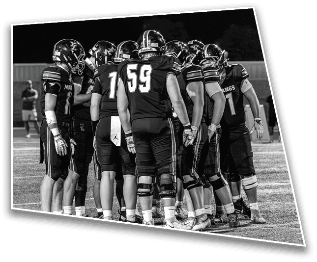 A group of football players in uniform and helmets are huddled together on a field during a nighttime game. Description generated by AI