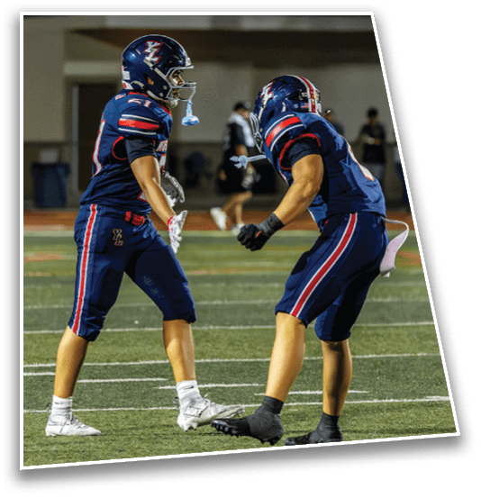 Two people in blue football uniforms with helmets are on a field, appearing to be engaged in a game or practice. Description generated by AI