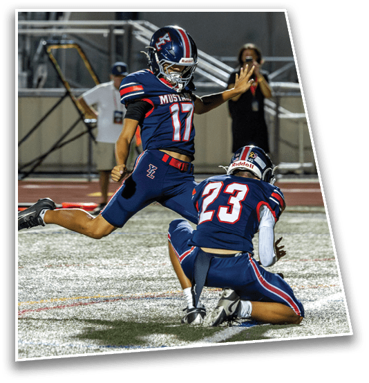 Two people in football uniforms are on a field with one person holding the ball on the ground while the other is kicking it. Description generated by AI