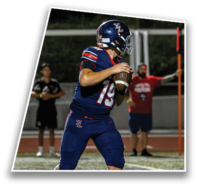 A person in a blue football uniform and helmet is holding a football and preparing to throw it on a field with two blurred people in the background. Description generated by AI