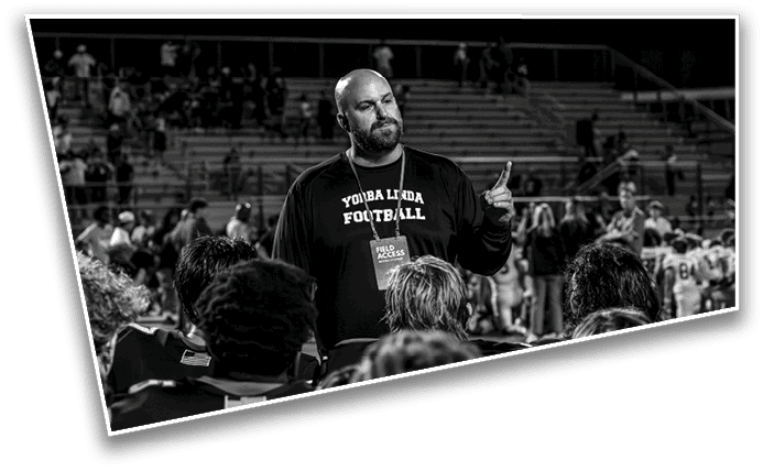 A person wearing a “Youba Linda Football" shirt is speaking to a group of people on a sports field with bleachers and spectators in the background at night. Description generated by AI