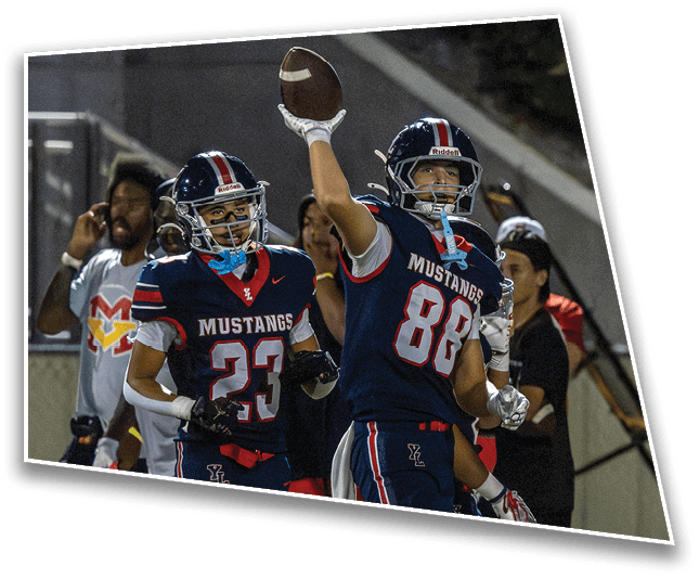 Two people in football uniforms and helmets celebrate on a field with one holding a football raised in the air. Description generated by AI