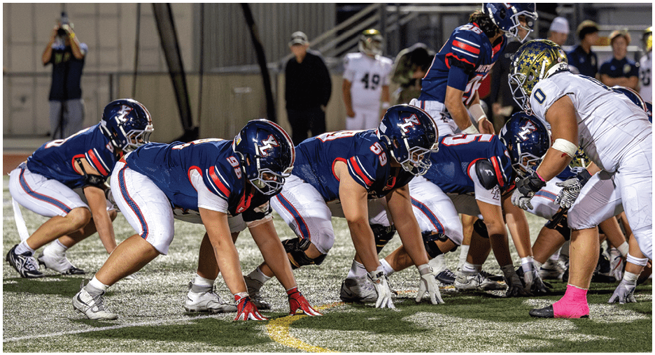A group of football players in blue uniforms are lined up in a crouched position facing players in white uniforms on a field during a game. Description generated by AI