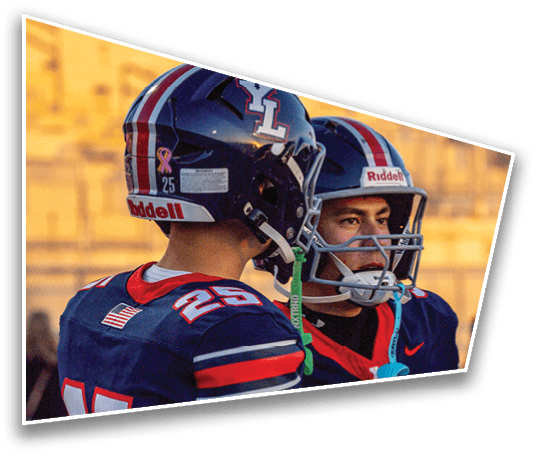 Two people in football uniforms and helmets walk on a field with other players and spectators in the background during a clear evening. Description generated by AI