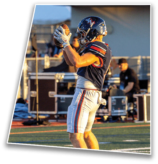 An adult wearing a football uniform and helmet is catching a football on a field with equipment and another person in the background. Description generated by AI