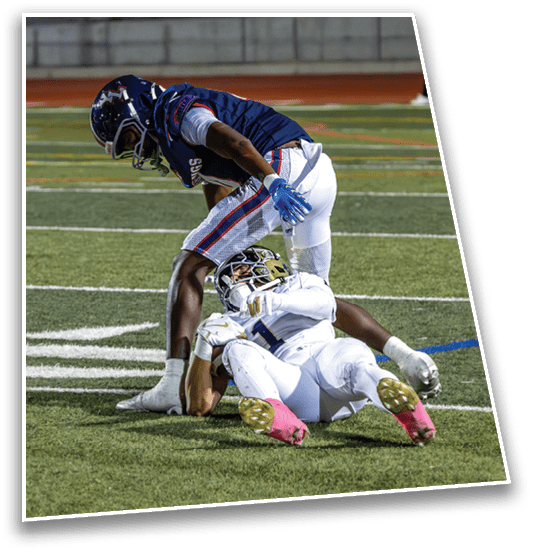 Two people in football uniforms are on a field with one person standing over another who is lying on the ground. Description generated by AI