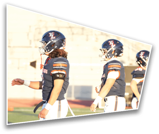 Two people wearing dark blue football uniforms and helmets with a red and white logo are walking side by side on a field with blurred bleachers in the background. Description generated by AI