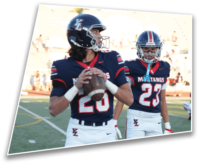 Two people wearing football uniforms and helmets stand on a field with one holding a football in a ready-to-throw position. Description generated by AI